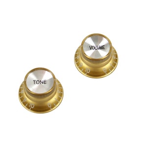 Peut inclure: Deux boutons de guitare dorés avec des chiffres et des lettres blancs. Le bouton de gauche indique "TONE" et le bouton de droite indique "VOLUME".