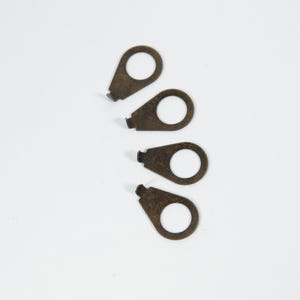 May include: Four small, bronze-colored metal tags with a teardrop shape and a circular hole. Each tag has a small rectangular tab at the narrow end. The tags are arranged on a white background.