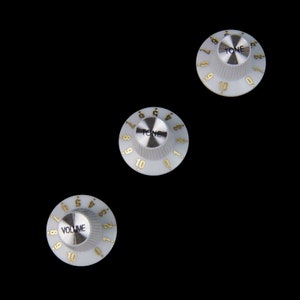 Pode incluir: Três botões de guitarra brancos com números e letras dourados. Os botões são etiquetados como "TONE" e "VOLUME".