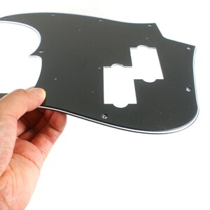 Puede incluir: Un golpeador de guitarra negro con un borde blanco, sostenido por una mano. El golpeador tiene dos recortes rectangulares y varios agujeros para tornillos. Es una pieza de repuesto para una guitarra.