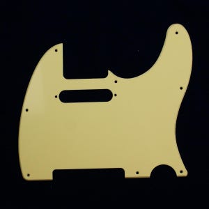 Pode incluir: Um pickguard de guitarra creme com uma forma distinta, apresentando um recorte retangular e vários orifícios para parafusos. O pickguard tem uma superfície lisa e sólida e é projetado para um modelo de guitarra específico.