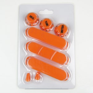 Puede incluir: Perillas de control y puntas de interruptor de guitarra naranja en un paquete de plástico transparente. Las perillas están etiquetadas como "TONE" y "VOLUME". El paquete incluye tres puntas de interruptor y tres cubiertas de interruptor naranjas.