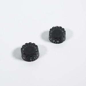 Könnte beinhalten: Zwei schwarze runde Knöpfe mit den Zahlen 0-10 darauf gedruckt. Die Knöpfe werden zum Stimmen von Musikinstrumenten verwendet.