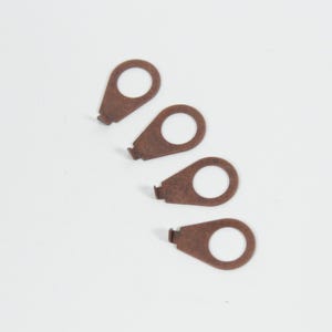 May include: Four small, copper-colored metal pieces with a circular hole and a small rectangular tab at the base. The pieces are arranged diagonally on a white background.