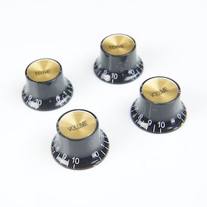 Op de afbeelding: Vier zwarte gitaarknoppen met gouden toppen. Twee knoppen zijn gemarkeerd met "TONE" en twee met "VOLUME". Elke knop heeft een genummerde schaal van 0 tot 10. De knoppen staan op een witte achtergrond.