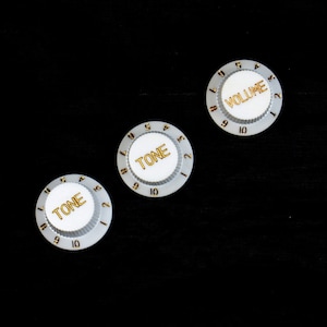 Pode incluir: Três botões de guitarra brancos com letras douradas. Os botões são etiquetados como "Volume", "Tono" e "Tono".