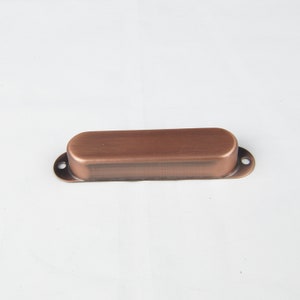 May include: A copper-colored metal cover for a guitar pickup. The cover is oval-shaped and has rounded edges.