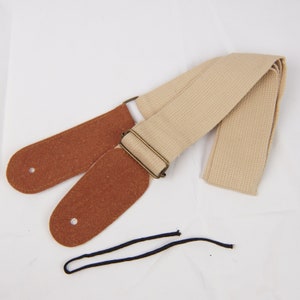 May include: A beige guitar strap with brown leather ends and a metal buckle. The strap is made of woven fabric and has a simple design.