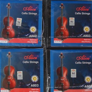May include: Four cello string packages are displayed, each with a cello illustration. The packages are blue with the "Alice Cello Strings" logo. Each package has the string type "A803 Steel Core Alloy Winding" and the string size, such as "4/4 A1".