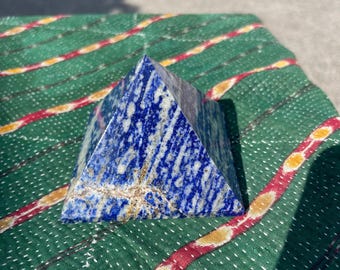 May include: A blue and white lapis lazuli pyramid. The pyramid is a deep blue colour with white and gold veining. It is sitting on a green fabric with red and yellow stripes.