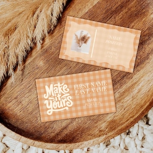 May include: Two peach-colored business cards with a gingham pattern and decorative edges rest on a wooden tray. One card features the text "Make it Yours" in a script font. The other card has a floral design and space for contact information.