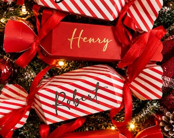 May include: Four rectangular gift boxes shaped like candy crackers, tied with red ribbons at both ends. Two boxes feature red and white diagonal stripes with the names John and Robert in black script. Two solid red boxes feature the names Henry and Rebecca.