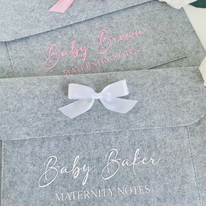 May include: Two grey felt envelopes with pink and white bows. The envelopes are labelled "Baby Brown Maternity Notes" and "Baby Baker Maternity Notes".