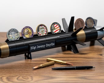 May include: A black, gold, and silver missile-shaped display stand holding several challenge coins. The stand has fins and the text "SSgt Jeremy Smythe". Two bullets and a pen are on the wooden surface.