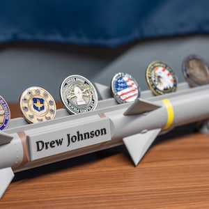 May include: A gray missile model with a yellow stripe and the name "Drew Johnson" engraved on the side. Six different military challenge coins are displayed in slots on the model.
