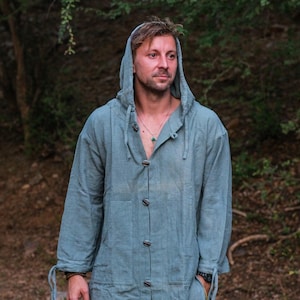 May include: A light blue hooded jacket with wooden buttons. The jacket has long sleeves with tie details at the wrists. The person in the image is wearing a necklace. The background is a blurred forest scene.