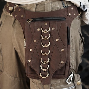 May include: A brown hip bag with a central pocket featuring decorative metal rings and rivets. The bag has a zippered main compartment and a smaller front pocket. The bag is attached to a brown belt and is worn around the waist.
