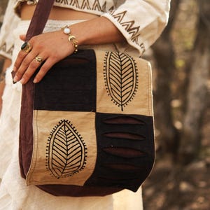 May include: A rectangular crossbody bag featuring a patchwork design of dark brown and tan fabric panels. The tan panels are decorated with a black leaf print, while the dark brown panels feature horizontal cut-out slits across the surface.