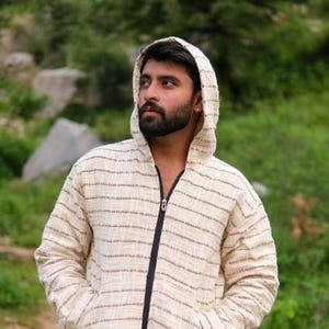 Könnte beinhalten: Cremefarbene Kapuzenjacke mit dünnen braunen horizontalen Streifen. Die Jacke hat einen durchgehenden schwarzen Reißverschluss und eine Kapuze. Der Stoff scheint gewebt zu sein. Die Person trägt die Jacke im Freien.