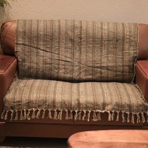 May include: A brown and gray striped throw blanket with fringe detail draped over a brown leather sofa. The blanket has vertical stripes and a textured appearance, adding a touch of rustic charm to the living space.