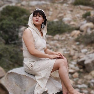 May include: A sleeveless, cream-colored hooded dress featuring a textured fabric, a V-neckline, and a flowing skirt that falls to the mid-calf. The garment includes a hood that rests over the head and shoulders, providing a simple, uniform appearance.
