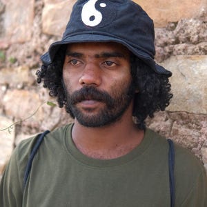 May include: Black bucket hat with a white yin-yang symbol. The hat is worn by a person wearing an olive green t-shirt. The background is a stone wall.