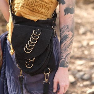 May include: A black canvas crossbody bag featuring a flap closure adorned with a vertical row of six brass-toned metal rings. The bag includes adjustable black straps with brass-toned lobster claw clasps and additional metal ring hardware details.