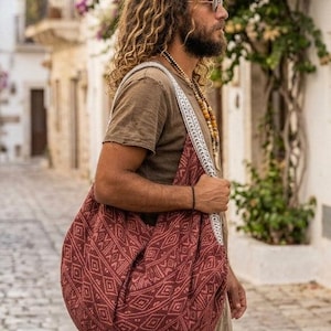 Könnte beinhalten: Eine große, weiche Hobo-Umhängetasche aus dunkelrotem Stoff mit einem sich wiederholenden weißen geometrischen Muster. Die Tasche hat einen breiten, integrierten Riemen mit einem komplexen weiß-grauen Webmuster, der über der Schulter liegt.