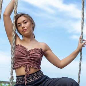 Könnte beinhalten: Ein mauvefarbenes, trägerloses Crop-Top mit Rüschenkante und Kordelzugdetail. Das Top wird mit einer schwarzen Hose mit besticktem Bund kombiniert. Der Hintergrund ist ein blauer Himmel mit Wolken.