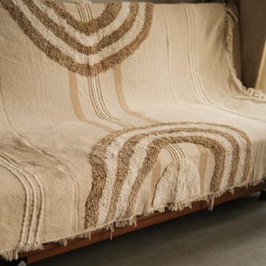 Op de afbeelding: Beige en bruine geweven sprei met een getuft regenboogontwerp. De deken heeft een gefranjerde rand en verticale strepen. De sprei is over een houten bank gedrapeerd, wat een gezellige en gestructureerde uitstraling creëert.