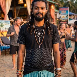 Puede incluir: Una persona con una camiseta negra con un patrón de ondas y pantalones azules con cordón. Está adornada con múltiples collares y pulseras de cuentas. El fondo muestra una escena de playa con otras personas y un letrero.