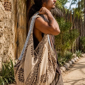 Puede incluir: Un bolso de hombro grande y holgado hecho de tela beige con un patrón geométrico marrón oscuro. El bolso tiene una correa ancha integrada con un detalle de borde negro y blanco estilo chevrón, llevado sobre el hombro.