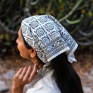 May include: A white bandana with a black spiral and geometric pattern. The bandana is tied around the head. The background is blurred, with green and brown tones.