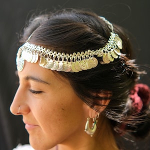 Puede incluir: Una diadema plateada adornada con colgantes en forma de monedas. La diadema se lleva en la cabeza y se combina con pendientes de aro plateados. La imagen es un primer plano que muestra los detalles de la joyería.