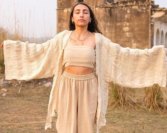 Puede incluir: Conjunto de tres piezas de lino beige que consta de un top de tubo, pantalones de pernera ancha y una chaqueta larga tipo kimono abierta. El conjunto está hecho de una tela ligera y texturizada. La modelo lleva collares superpuestos y pendientes.