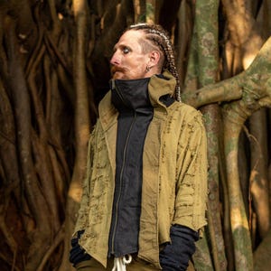 Könnte beinhalten: Eine tanfarbene Jacke mit einer schwarzen Weste mit Reißverschluss darunter. Die Jacke hat ein abgenutztes, strukturiertes Aussehen. Die Person trägt schwarze Handschuhe und hat geflochtenes Haar. Der Hintergrund zeigt einen großen Baumstamm.