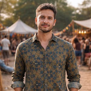 Puede incluir: Un hombre con una camisa de manga larga con un estampado floral azul y dorado repetido. La camisa tiene cuello y mangas enrolladas. En el fondo se ve una multitud y tiendas de campaña.