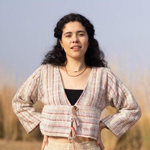 Può includere: Top a righe beige, rosse e bianche con scollo a V e chiusura con lacci. Il top ha maniche lunghe e viene indossato sopra un top nero. La modella indossa una collana d'oro.