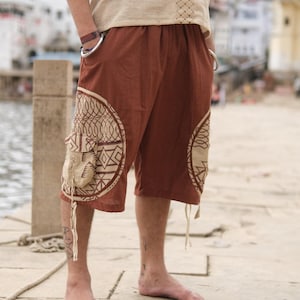 May include: Knee-length brown shorts featuring large circular beige side panels with dark brown geometric patterns. The shorts include side cargo pockets with flaps and thin drawstring ties hanging from the bottom edges of the pockets.