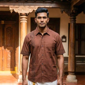 May include: A short-sleeved, brown button-down shirt with a decorative pattern on the left side and sleeve cuffs. The shirt has a chest pocket and is worn with white pants. The background features wooden architectural elements.