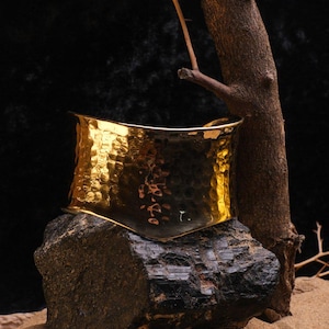 May include: A wide, hammered gold-tone cuff bracelet. The bracelet has a textured surface and a curved shape. It is displayed on a dark rock with a black background and a small tree branch.