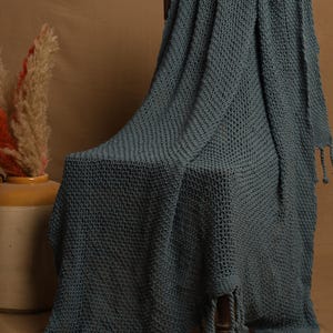 May include: A teal-colored knitted throw blanket draped over a wooden chair. The blanket has a textured weave and knotted fringe along the edges. The background features a neutral-toned backdrop and a decorative vase with dried plants.