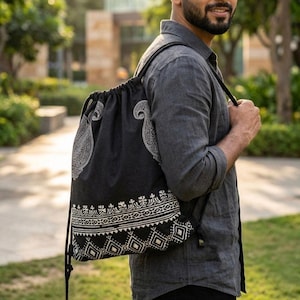 Könnte beinhalten: Schwarzer Rucksack mit Kordelzug und weißen Paisley- und geometrischen Mustern. Die Tasche hat einen Kordelzugverschluss und Schultergurte. Das Design umfasst ein großes Paisley-Motiv und einen dekorativen Rand. Die Tasche ist aus dunklem Stoff.