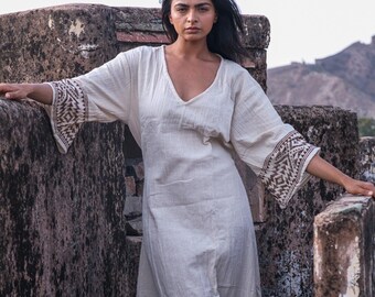 May include: A cream-colored, V-neck dress with wide sleeves featuring a geometric brown pattern on the cuffs. The dress has a relaxed fit and is made of a lightweight fabric. The model is standing on a stone wall.