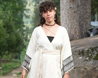 May include: A cream-colored wrap top with black and white geometric patterned sleeves and a matching belt. The top is paired with beige pants. The person is wearing a black top underneath, a necklace, and earrings. The background is a natural outdoor setting.