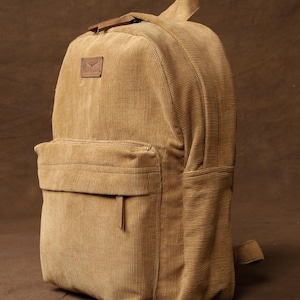 Może przedstawiać: Brązowy plecak z  cordury z brązową skórzaną naszywką z napisem "Travelsmith". Plecak ma kieszeń z zamkiem błyskawicznym z przodu.
