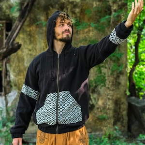 Puede incluir: Chaqueta negra con capucha y cremallera con detalles geométricos blancos en las mangas y bolsillos. La chaqueta se combina con pantalones color canela con un diseño de símbolo de mano. El conjunto es adecuado para uso casual.
