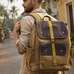 May include: A tan and brown canvas backpack with a flap closure and two front pockets. The backpack has a yellow strap down the centre and is worn by a person walking down a street.