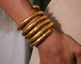 Puede incluir: Juego de pulsera y anillo de serpiente dorados. La pulsera se enrolla alrededor de la muñeca en un diseño en espiral, con una cabeza de serpiente en la parte superior. El anillo presenta un diseño enrollado similar. Las joyas se muestran en el brazo y la mano de una persona.