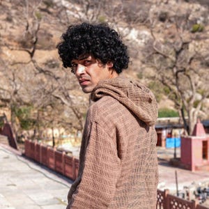 May include: A brown hooded top with a textured weave. The person wearing it has dark curly hair and is looking over their shoulder. The background shows a natural outdoor setting with trees and a stone structure.